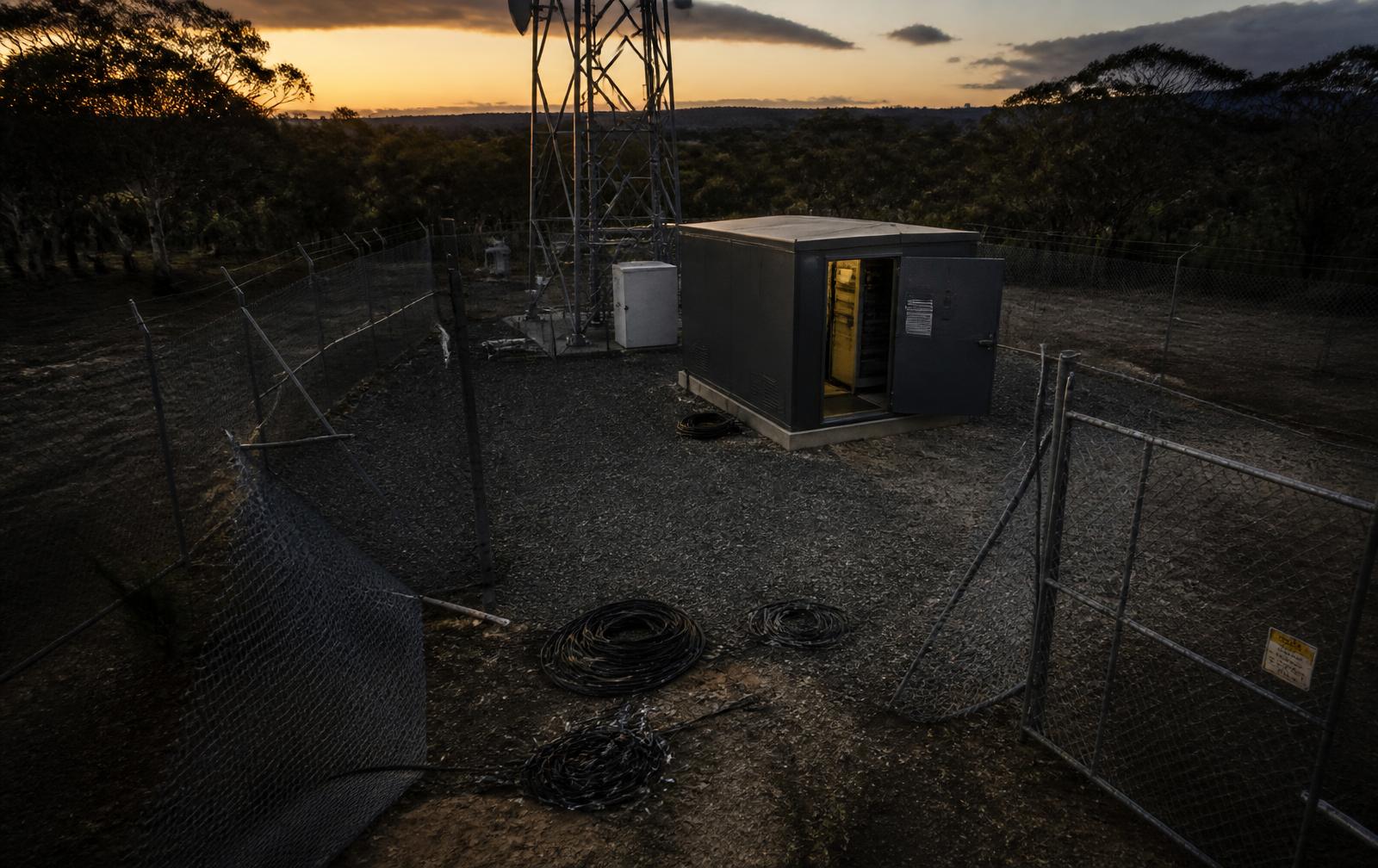 Aerial view of a remote telecommunications compound after a break-in, showing cut perimeter fencing, an open equipment shelter and stolen cabling on the gravel pad