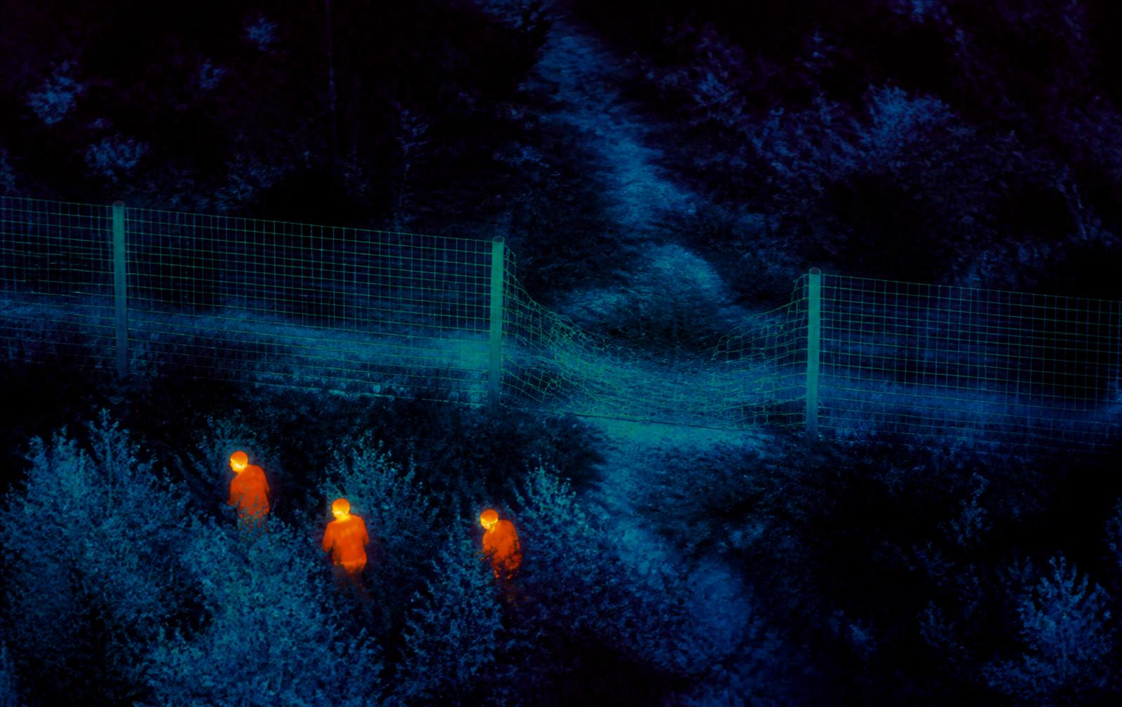 Thermal imaging aerial view showing a cut perimeter fence with heat signatures of people hiding behind bushes
